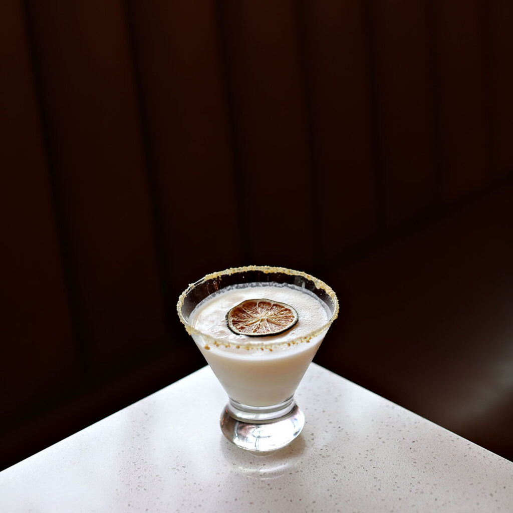 A fancy white drink sits on a white table against a dark brown backgrround