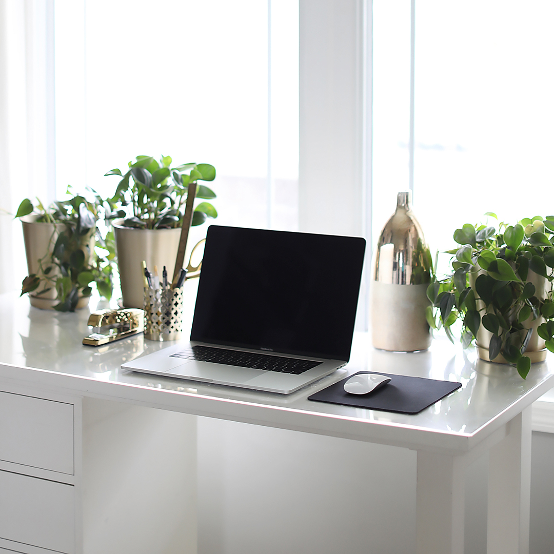 An open laptop computer sits on a white desk surrounded by green plans and gold desktop accessories